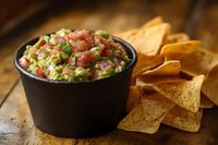 Fresh guacamole dip with diced tomatoes, red onions and cilantro served in black bowl with crispy corn tortilla chips on rustic wooden table background.