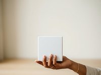 Hands holding blank white box on neutral beige background, suitable for product packaging mockups, branding presentations, and commercial advertising campaigns.