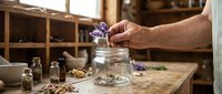 Adult hand placing dried purple flowers into glass jar with herbs and botanicals on wooden table in apothecary or wellness studio setting for natural remedy preparation.