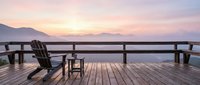 Wooden deck with Adirondack chairs overlooking calm lake at sunset, mountains in background. Serene waterfront retreat setting for relaxation and travel concepts.