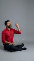Young man with dark hair and beard, South Asian ethnicity, wearing red button-up shirt and dark jeans, sitting cross-legged while holding laptop and pointing upward with inspiration. Isolated on gray background for web, app, and marketing use.