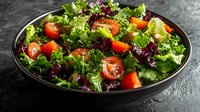 Fresh mixed greens salad with cherry tomatoes and colorful vegetables in black bowl on dark slate background for healthy eating and nutrition.