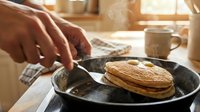 Hands adding butter to cast iron skillet with golden pancakes for breakfast cooking in warm kitchen sunlight.