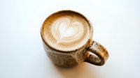 Overhead view of hot cappuccino with latte art heart design in ceramic mug on white surface, suitable for cafe menus and beverage marketing.