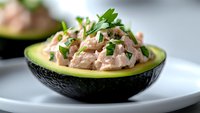 Fresh tuna salad served in ripe avocado half, garnished with chopped parsley on white plate, close-up view showing creamy texture and vibrant colors.