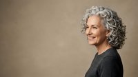 Mature woman with gray curly hair, Caucasian ethnicity, wearing dark sweater, smiling at camera against neutral beige background. Portrait for aging, beauty, confidence themes.