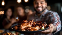 Hispanic man serving grilled chicken wings on black plate at dinner party with friends, warm ambient lighting and festive atmosphere in background.
