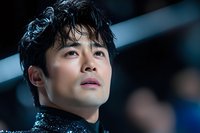 Young Asian male performer in sequined black costume gazing upward with dramatic lighting, expressing determination and artistry during stage performance.