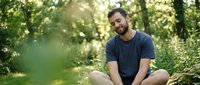 Man with dark beard and medium skin tone sitting in lush green forest, wearing casual navy t-shirt, smiling at camera during daytime. Suitable for lifestyle, wellness, nature, and outdoor activity content.