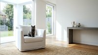 Modern minimalist living room interior with tabby cat sitting on white armchair near large windows overlooking garden, natural light, contemporary home decor.