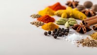 Colorful spices and herbs arranged on white surface with turmeric, paprika, cinnamon, cardamom, peppercorns creating aromatic culinary ingredient display.
