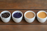 Row of white ceramic cups containing coffee beans, dried lavender, creamy coffee, and golden espresso on rustic wooden table background.