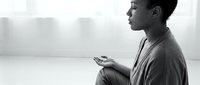 Young man of South Asian descent sitting in contemplative pose holding smartphone, looking away pensively in minimalist interior space with natural window light, suitable for mental health, technology addiction, communication, and lifestyle editorial use.