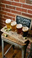 Craft beer flight featuring four glasses of varying shades displayed on rustic wooden board with hop garnish against brick wall and chalkboard sign for brewery tasting experience.