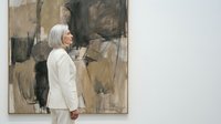 Middle-aged woman in white suit viewing abstract painting in modern art gallery. Professional visitor appreciating contemporary artwork in museum space.