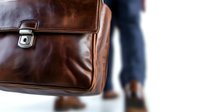 Brown leather briefcase with metal clasp in close-up view against blurred business person walking, showcasing professional accessory detail and craftsmanship.