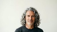 Mature man with long gray wavy hair and facial hair wearing dark casual shirt against neutral background, suitable for lifestyle and professional portraits.