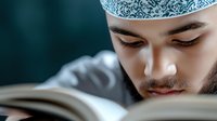 Young Middle Eastern man wearing traditional taqiyah cap reading religious book with focused expression against dark background, spiritual study concept.