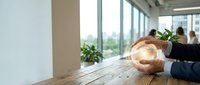 Businesswoman holding glowing light bulb at wooden desk in modern office with large windows, symbolizing innovation, creativity, and new business ideas for corporate success.