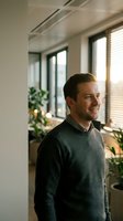Smiling Caucasian businessman in gray sweater standing in modern office with plants and natural light streaming through windows.