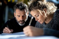 Middle-aged couple working together on paperwork at home, focused on signing documents. Woman in gray sweater and man with salt and pepper beard review contracts.