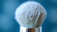 Macro shot of luxury shaving brush with white synthetic or natural bristles in metal holder against blurred blue background, highlighting grooming accessories and personal care.