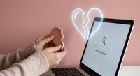 Young woman holding heart-shaped cookie while using laptop for online dating or social media connection on pink background for romantic digital lifestyle.