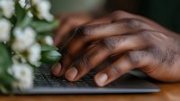 African American hands typing on black keyboard with white flowers and green leaves in soft focus background, creating peaceful work environment and wellness concept.