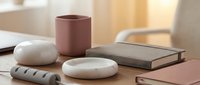 Minimalist workspace setup with pink ceramic mug, smooth white stones, gray notebook and modern accessories on wooden desk for productivity and zen office decor.