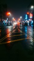 Wet city street at night with glowing traffic lights and neon signs reflecting on rain-soaked asphalt creating atmospheric urban scene with cars and pedestrians.