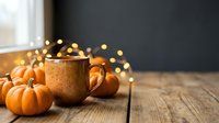 Warm mug with hot beverage surrounded by small decorative pumpkins and golden bokeh lights on wooden surface, autumn seasonal decoration for fall holiday marketing and seasonal content.