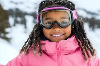 Cheerful African American girl wearing pink ski jacket and goggles with snow-covered hair smiles during winter mountain adventure. Active lifestyle and winter sports concept.