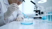 Scientist in white lab coat using pipette to drop blue liquid into petri dish during laboratory research experiment with microscope and test tubes.