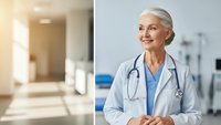 Senior female doctor with gray hair smiling confidently in white coat and stethoscope standing in modern hospital corridor with professional healthcare setting.