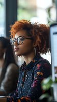 Young African American woman with natural curly auburn hair and glasses gazes thoughtfully in profile, wearing patterned blouse in modern office setting.