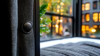 Cozy evening atmosphere with window view, warm bokeh lights outside, gray blanket on windowsill, black curtain detail in foreground creates intimate home setting.
