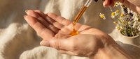 Natural skincare oil being dispensed from dropper bottle into cupped hands with dried flowers in soft sunlight for beauty and wellness routine.