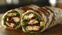 Grilled chicken wrap with fresh lettuce, avocado slices and drizzled sauce, rolled in flour tortilla, cut diagonally to show filling on wooden surface.