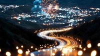 Glowing winding road at night with light trails and fireworks in sky above city lights, bokeh foreground creates magical atmosphere. Scenic mountain landscape.
