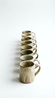 Row of handmade ceramic mugs with speckled glaze in earth tones arranged in line on white background for kitchen and dining design projects.