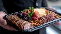 Gourmet meat and cheese platter with fresh berries, nuts and cured meats arranged on black catering tray, held by server in professional setting.