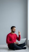 Young Indian man in red sweater sitting with laptop pointing up having eureka moment against gray wall for business presentations and startup concepts.