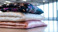 Stack of luxurious silk pillowcases in floral print, pink, white and navy blue colors arranged on reflective surface with soft natural lighting in bedroom setting.