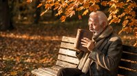 Elderly Caucasian man reading a book on a park bench surrounded by golden autumn leaves, enjoying a peaceful outdoor moment.