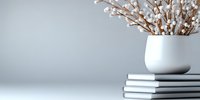 White ceramic vase with dried pussy willow branches on stack of books against neutral gray wall background, minimalist home decor composition with copy space.