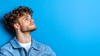 Young Caucasian man with curly hair and beard wearing denim jacket against vibrant blue background, eyes closed with contemplative expression. Copy space.