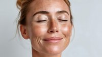Young woman with freckles and closed eyes smiling peacefully against light background, showcasing natural beauty and serene expression.