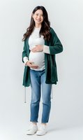 Young Asian pregnant woman in green cardigan and jeans smiling while holding belly on white background. Casual maternity fashion and lifestyle concept.