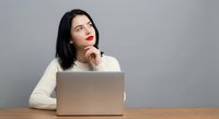 Young Asian businesswoman in white sweater thinking while working on laptop at wooden desk against gray background for office and workplace concepts.