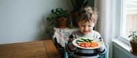 Young blonde Caucasian boy smiling while holding colorful plate of fresh vegetables in bright kitchen with plants and natural window light for healthy eating.